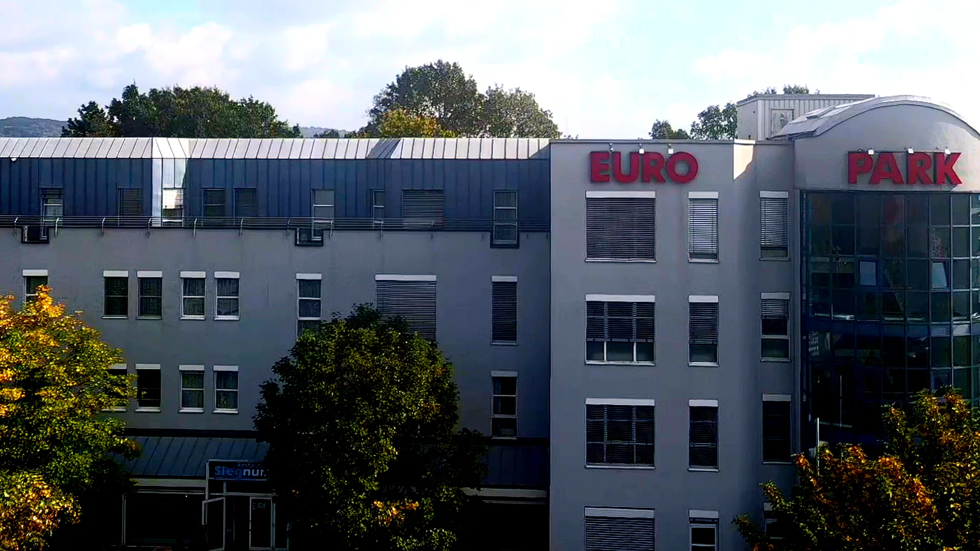 Drohnenaufnahme der Frontfassade des EURO PARK HOTEL Hennef: roter Schriftzug EURO PARK HOTEL auf dem Dach, verglaster Treppenturm über dem Eingang, Bäume im Vordergrund, darüber ein Himmel mit Wolken