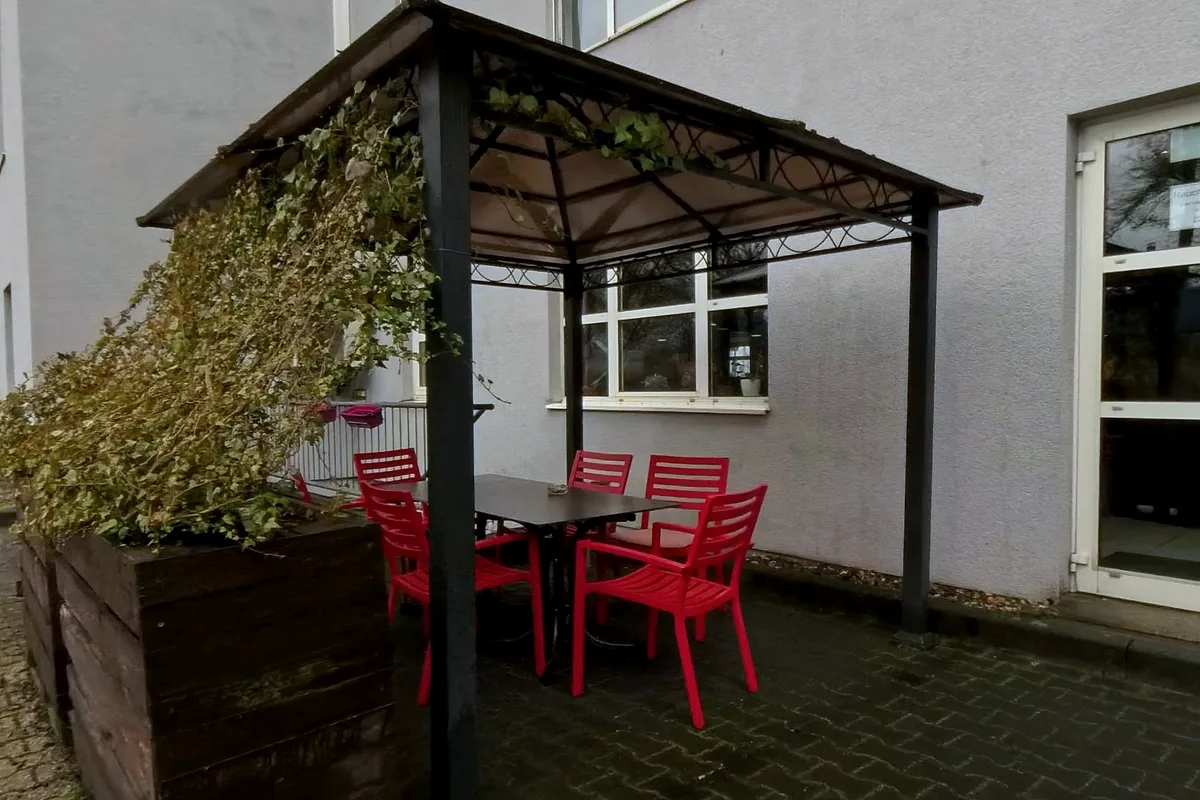 Terrasse an der Rückseite des EURO PARK HOTEL Hennef: Pavillon mit Efeu, roten Stühlen und dunklem Tisch auf gepflastertem Boden neben der Hausfassade